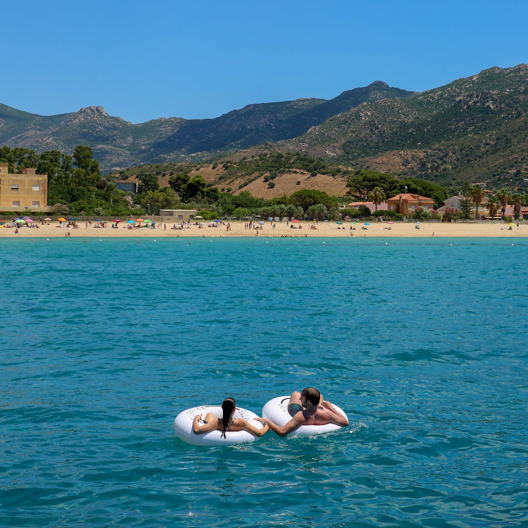 Man and woman floating far from the shore, looking back at the shore. The swim rings are white loteli swim rings. 