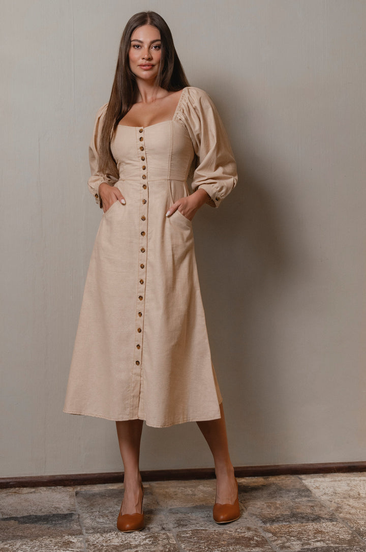 Woman wearing a beige linen dress with buttons standing against a plain wall.