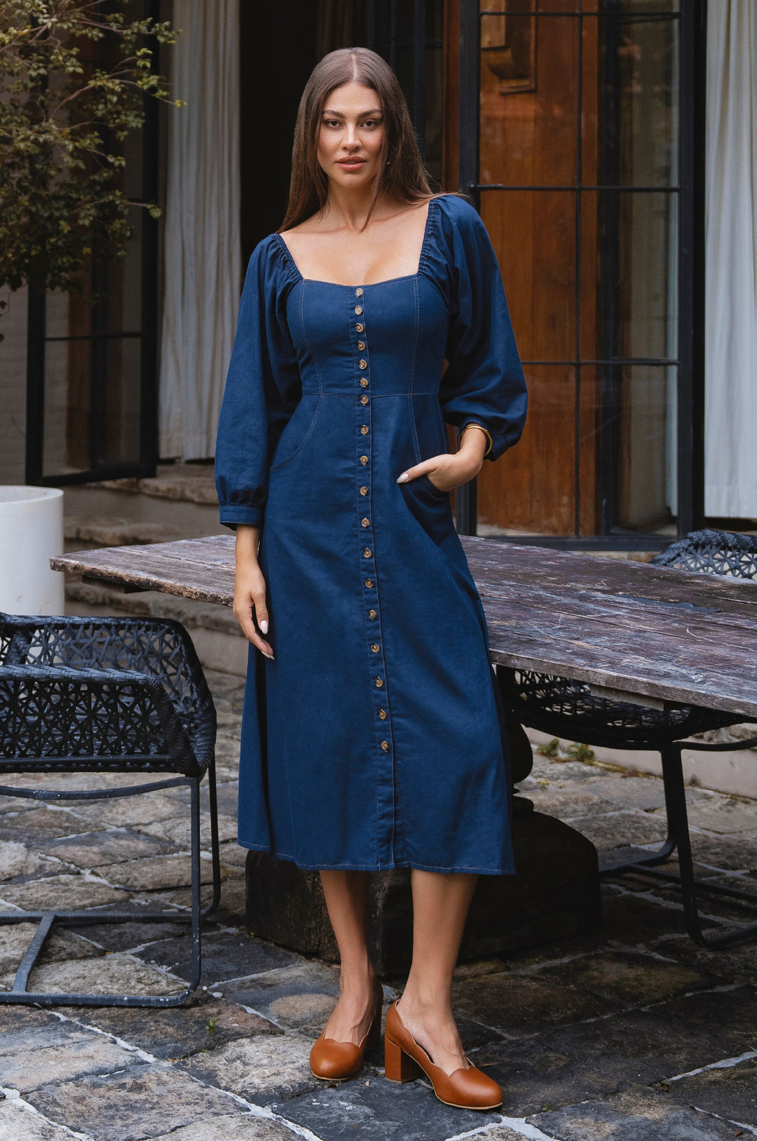 Woman wearing a blue linen dress standing outdoors on a stone patio.