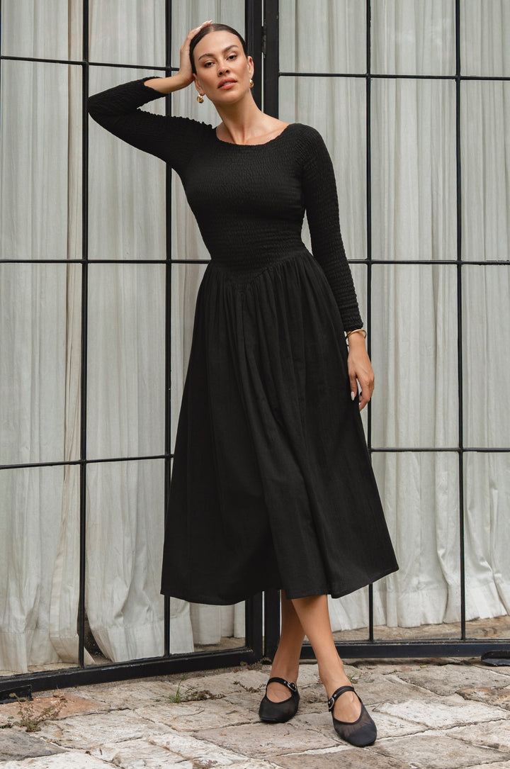 Woman wearing a black smocked dress standing against a textured wall.