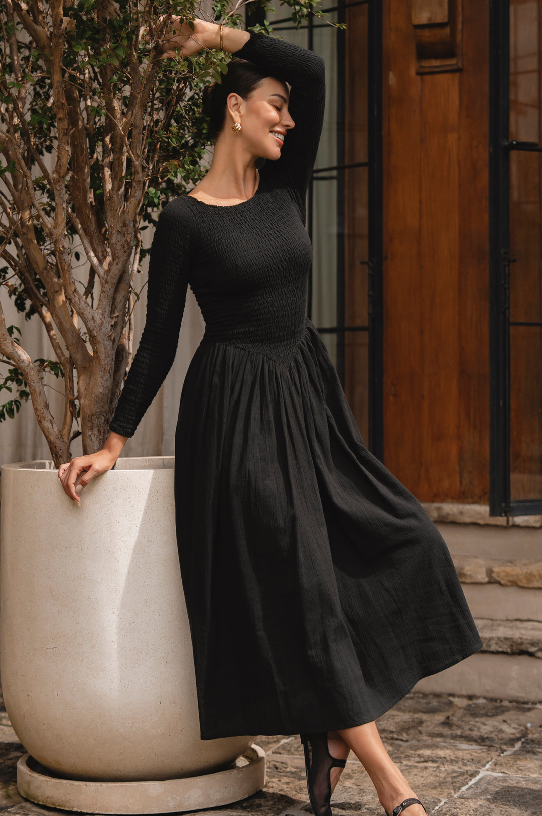 Woman in a black linen smocked dress standing next to a large planter outdoors.