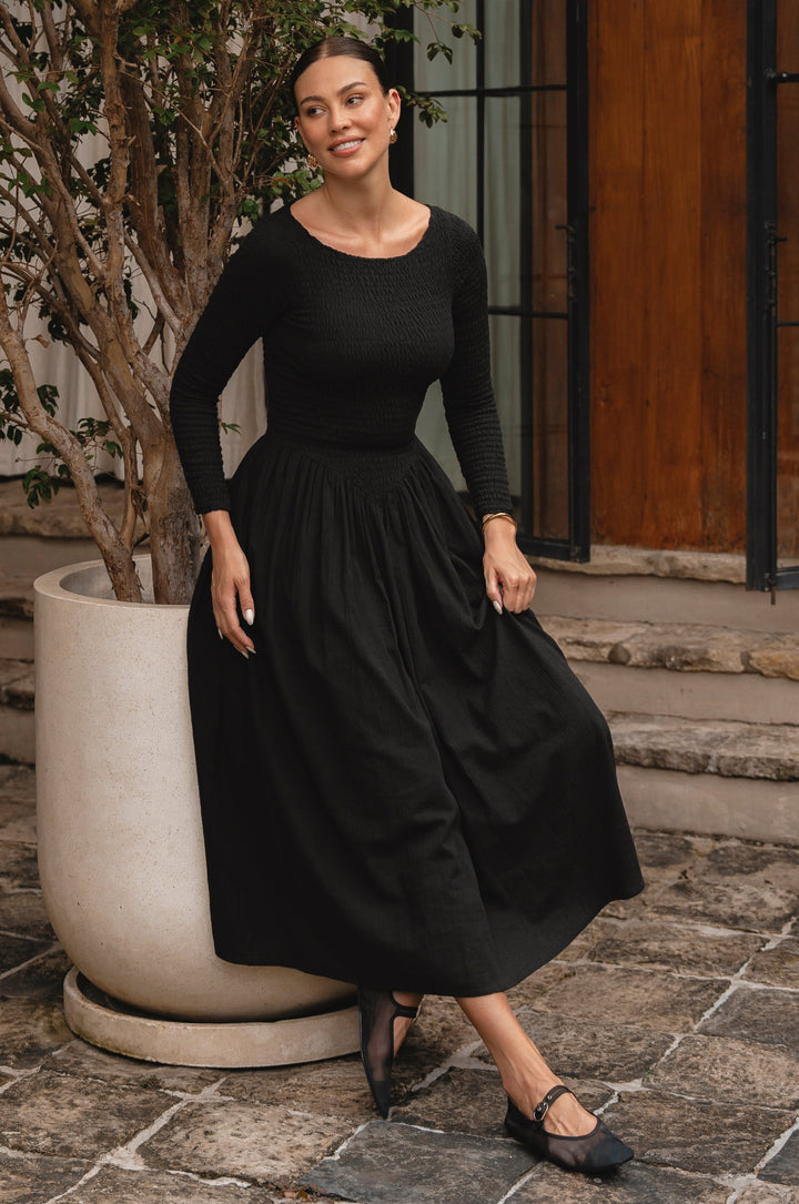 Woman wearing a black smocked dress standing outdoors on stone steps.