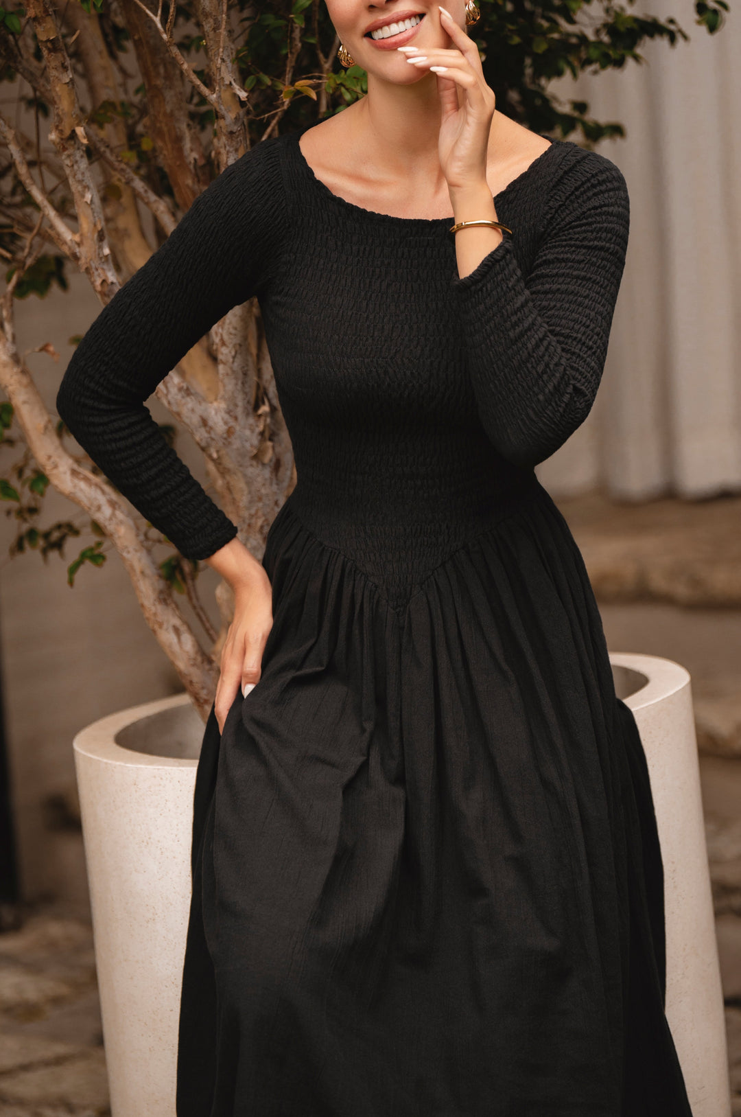 Woman wearing a black midi dress standing next to a decorative plant indoors.