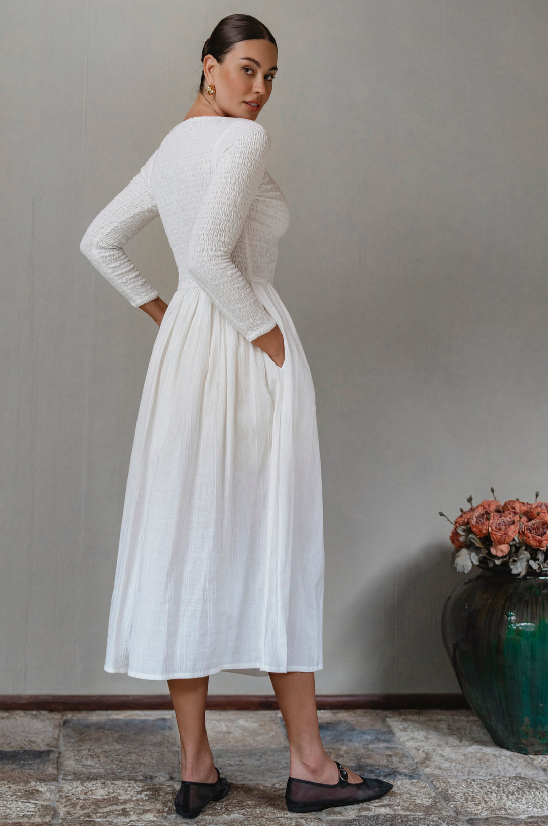 Woman wearing a white smocked long sleeve midi dress standing against a plain wall with a vase of flowers in the background.