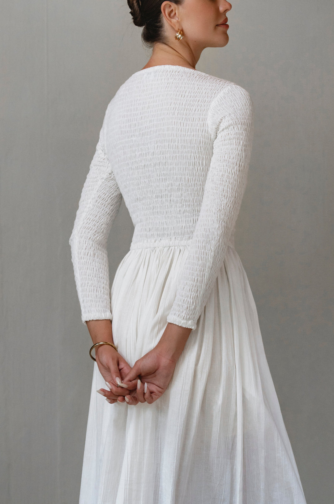 Woman wearing a white smocked long sleeve midi dress with long sleeves against a gray background