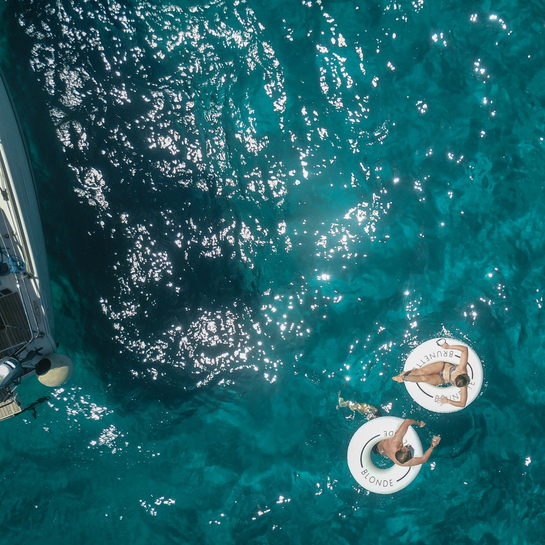 Aerial photo of a man and woman in white pool rings in the ocean near a boat. The man's swim ring has the word Blonde. The woman's swim ring has the word brunette on it.