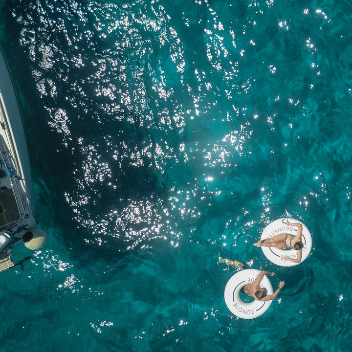 Aerial photo of a man and woman in white pool rings in the ocean near a boat. The man's swim ring has the word Blonde. The woman's swim ring has the word brunette on it.