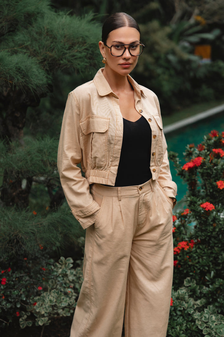 Woman wearing a beige jacket and pants with a black top, standing outdoors with greenery and flowers in the background- cottage core style