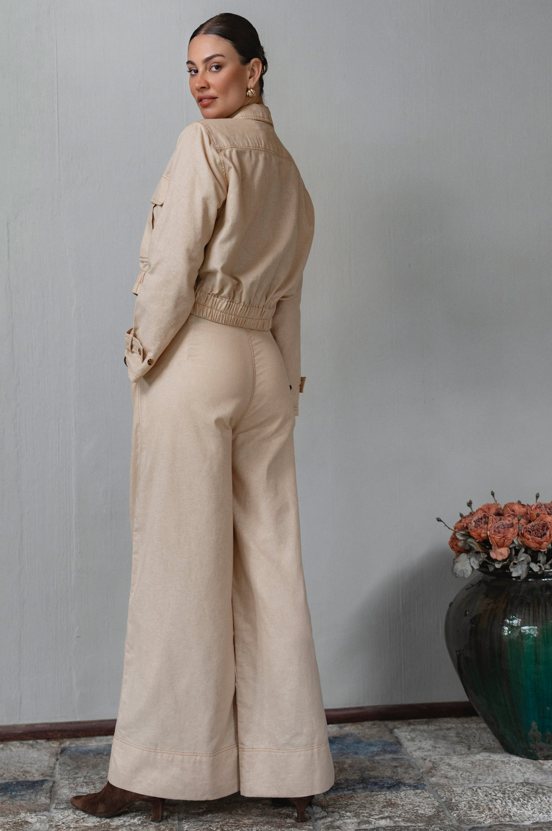 Woman wearing a beige outfit standing in a room with a vase of flowers on a table.