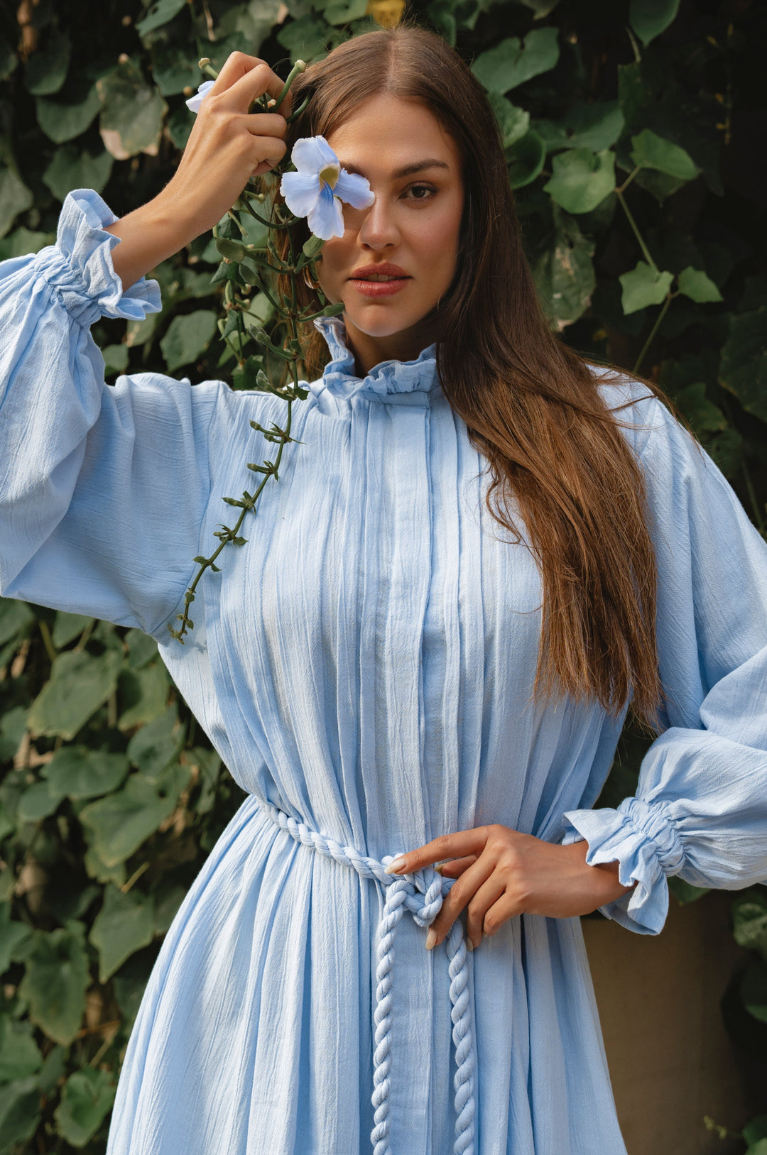 Woman in a light blue dress holding a flower in front of her face with a green leafy background