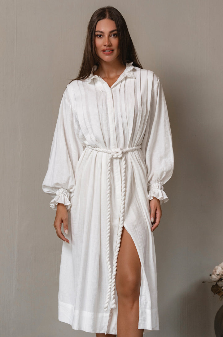 Woman wearing a white linen midi dress with a belt and slit on a plain background